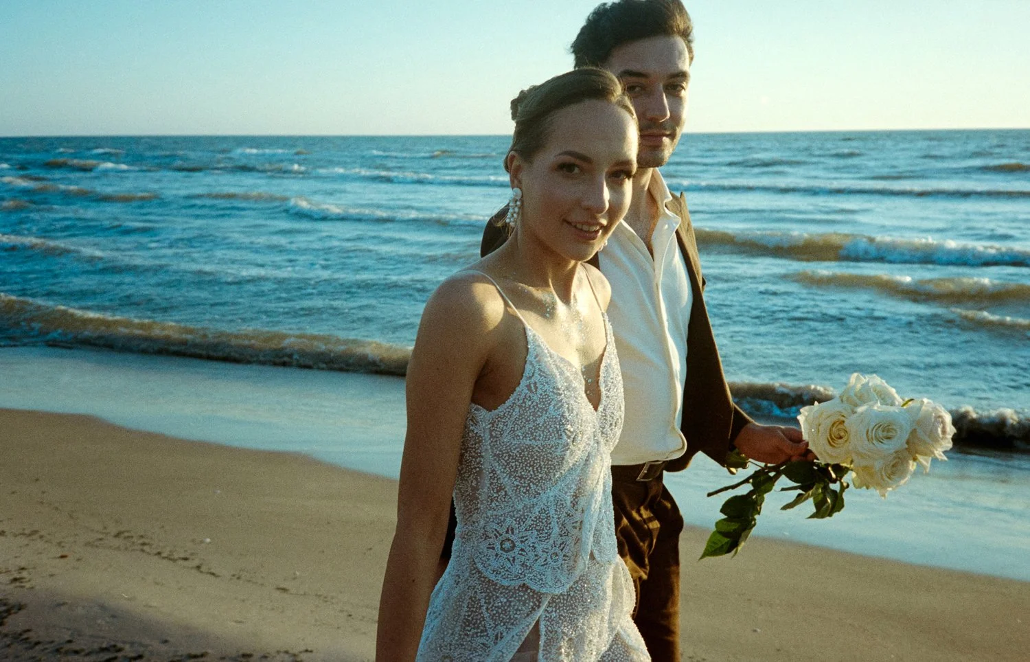 A newlywed couple walking on the beach during sunset, with the woman in a white lace wedding dress and the man in a white shirt and brown jacket holding a bouquet of white roses.