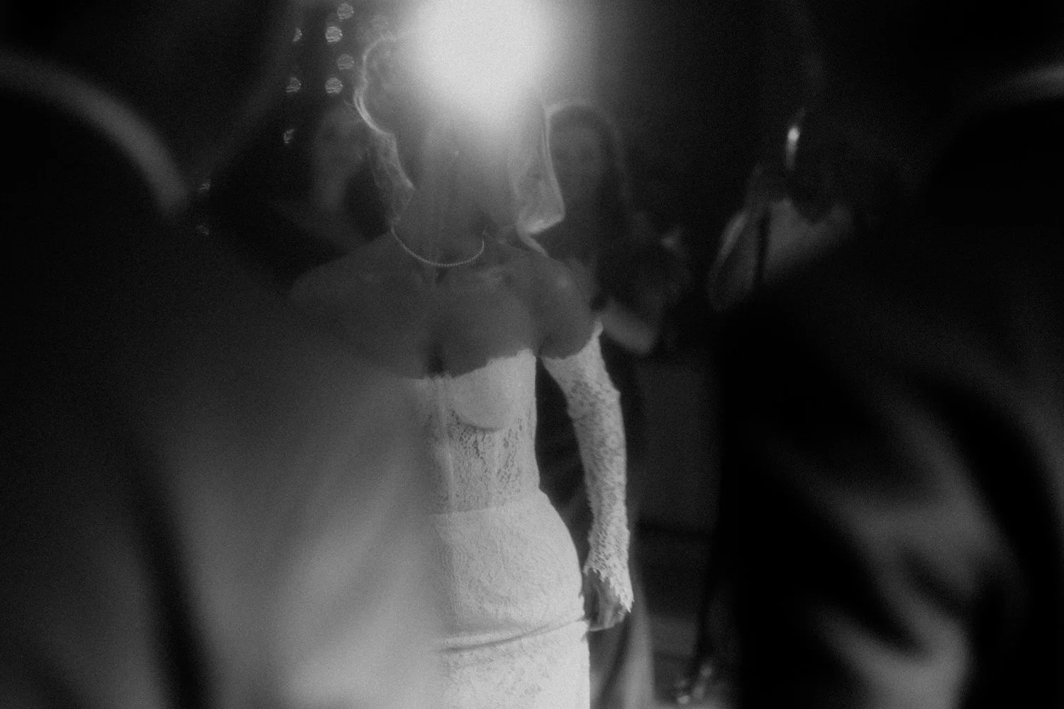 Black and white photo of a woman in a dress with lace sleeves and a pearl necklace, facing slightly downward, with super bright light behind her head.
