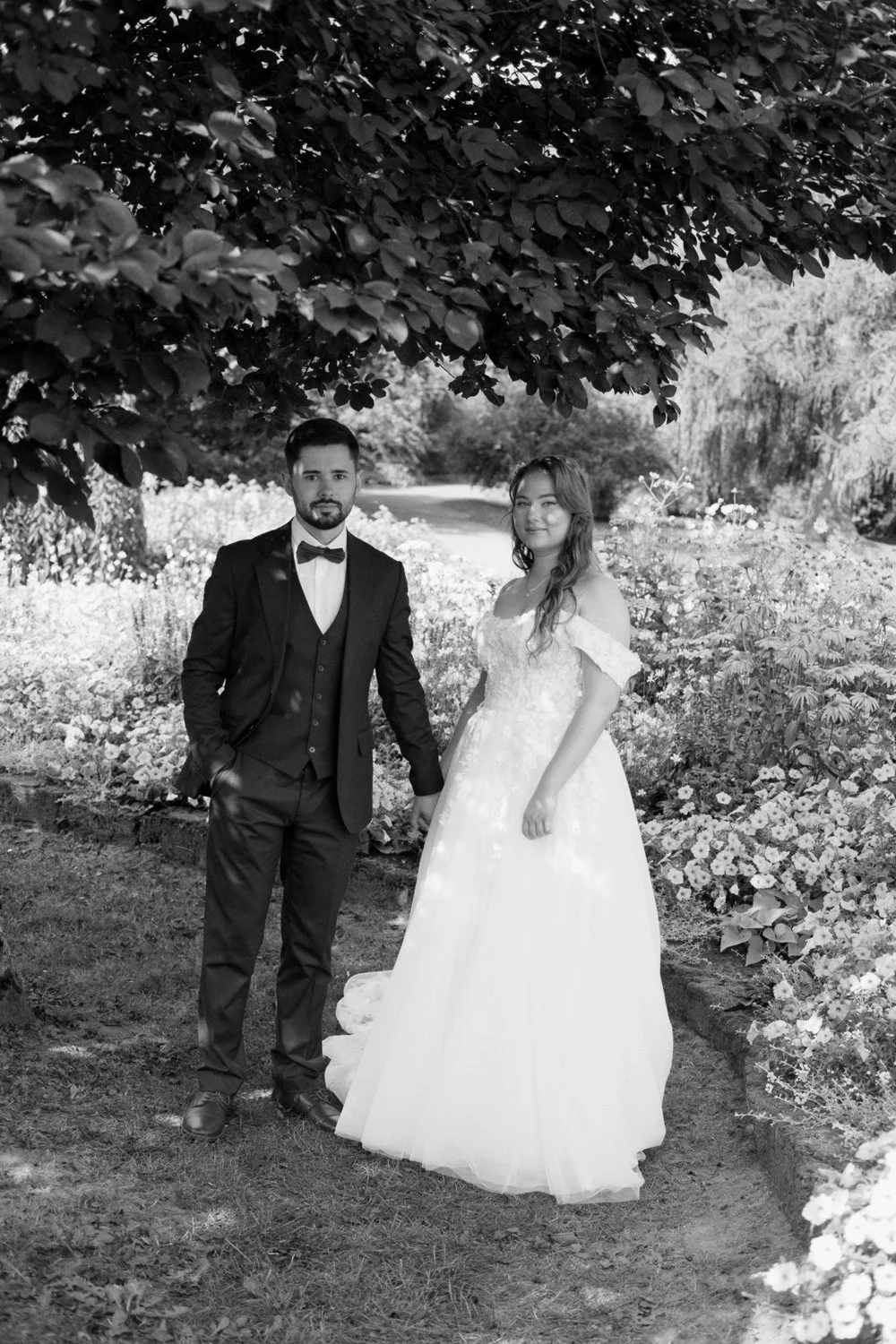 A black and white photo of a newlywed couple standing outdoors under a large leafy tree, with a garden of flowers in the background. The groom is dressed in a tuxedo with a bow tie, and the bride is in a white wedding gown with lace details.