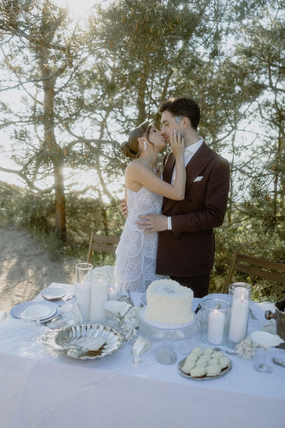 A bride and groom are sharing a kiss during their outdoor wedding reception, surrounded by trees. The table in front of them is decorated with a white cake, candles, and plates.