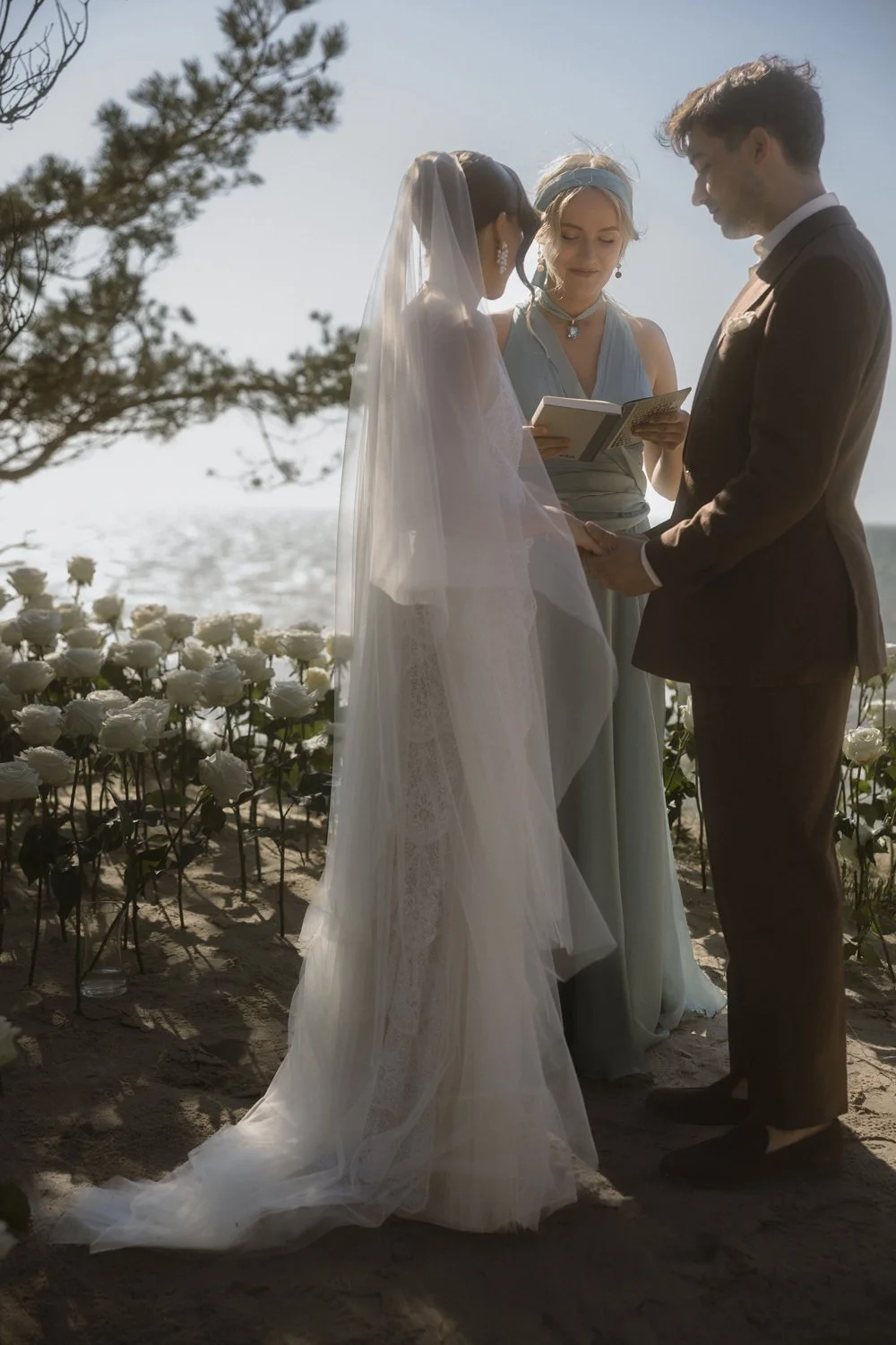 Bride and groom holding hands during their beach wedding ceremony, with officiant reading vows, surrounded by white roses and sand, seaside background with trees and ocean.