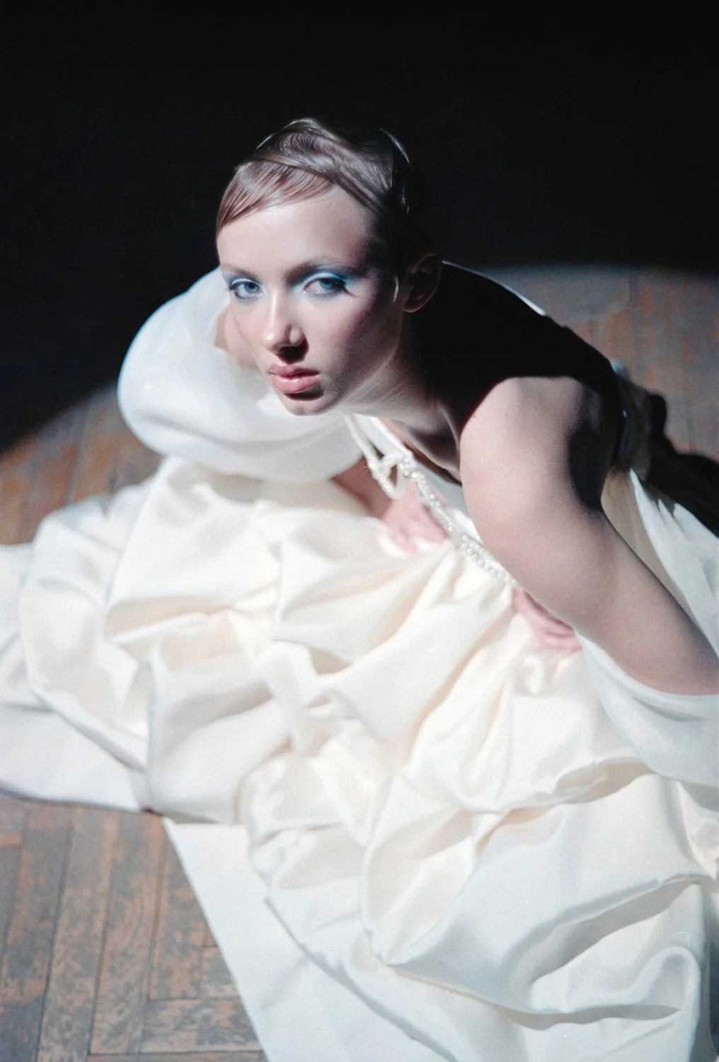 A woman in a white gown with a black stripe and silver embellishments sitting on a wooden floor, looking up at the camera with blue makeup and slicked-back hair.