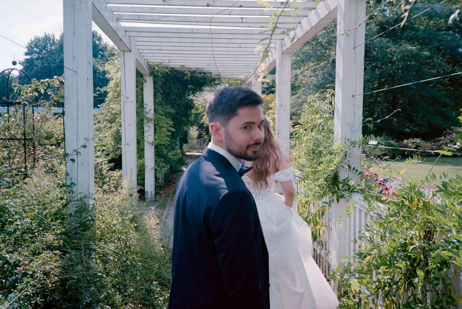 A man in a tuxedo stands in front of a woman in a white wedding dress, outdoors in a garden with green foliage and a wooden trellis structure.