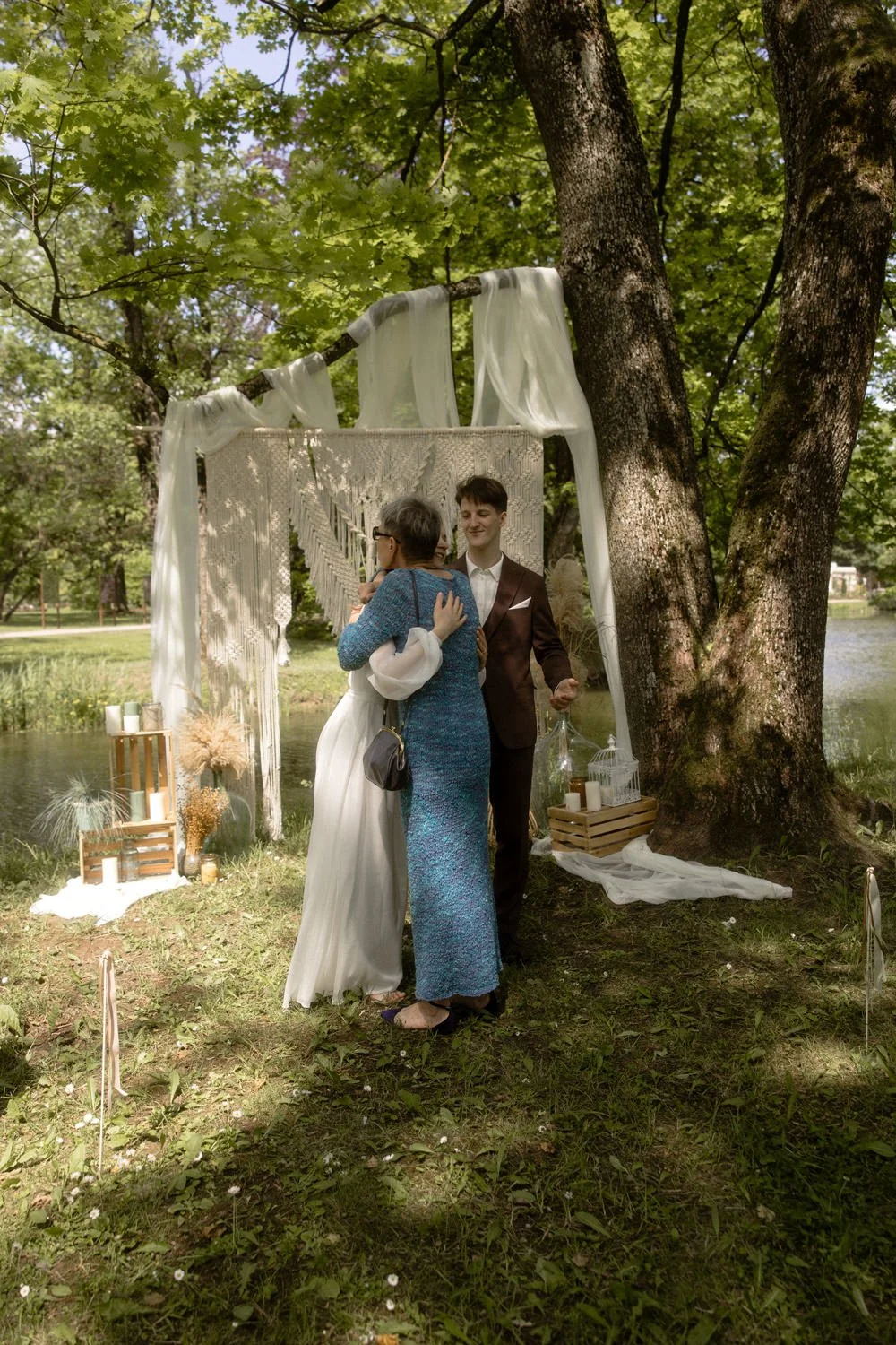 Couple in wedding attire sharing a hug beneath decorative arch in a lush outdoor setting near a river, with candles and decorative elements nearby.