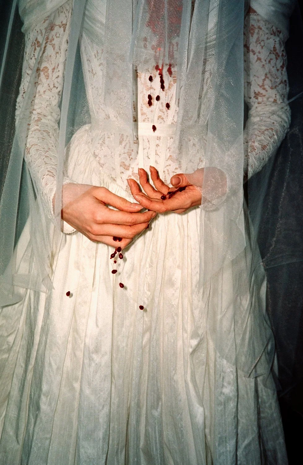 Close-up of a woman in a white lace dress with bloodstains and drops of blood on her hands and dress, holding small red seeds or berries.