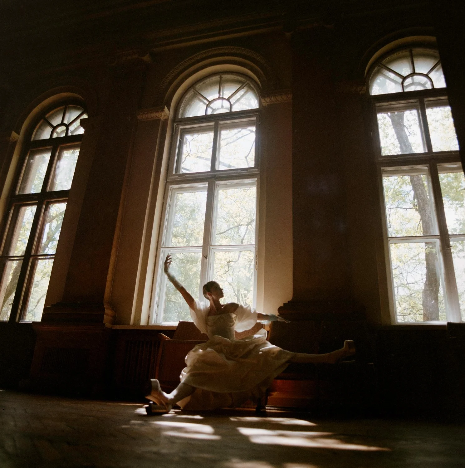 A ballet dancer in a white tutu performs a dance in front of tall arched windows with natural light streaming in, creating silhouette and shadow on the floor.