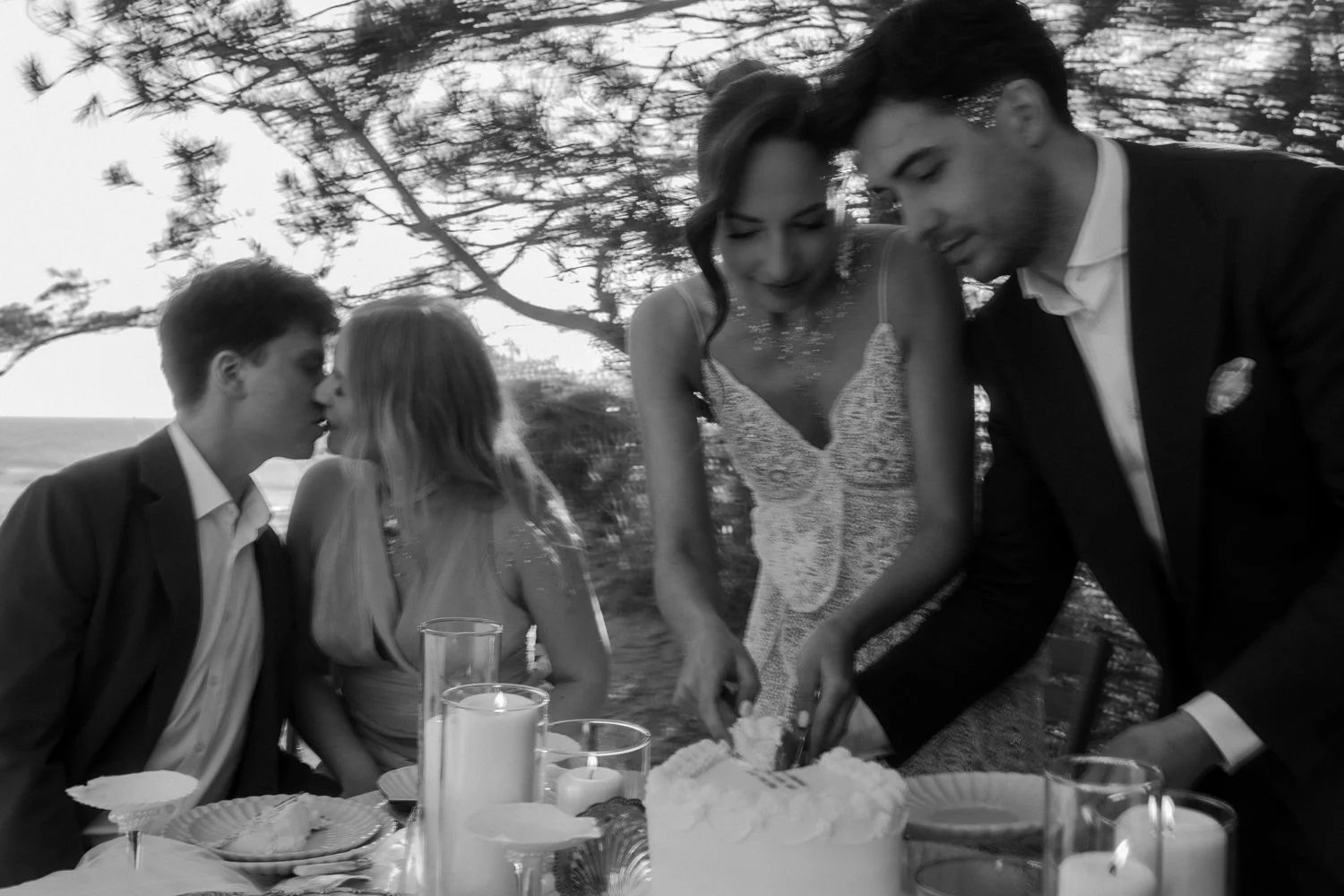 Four people at elopement, two men and two women, with a cake and candles on the table, outdoors near trees, in black and white.