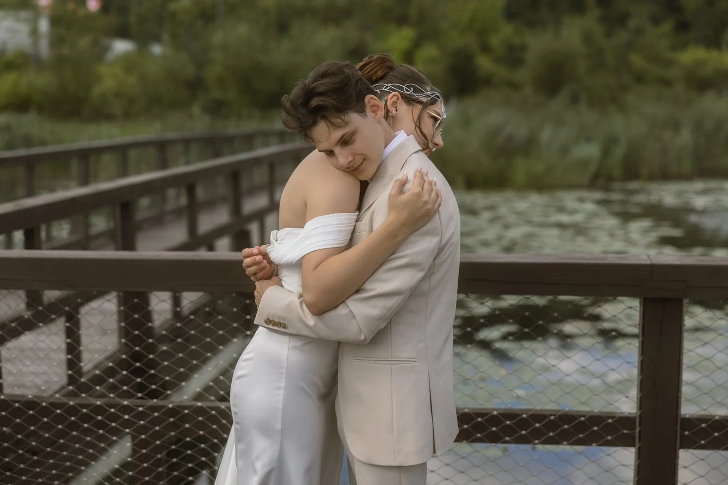 A couple embraces on a wooden bridge over a body of water, surrounded by lush greenery.