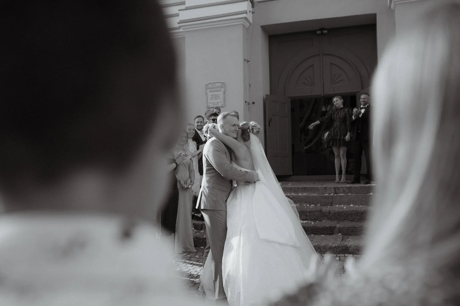 A black and white photo of a newlywed couple sharing a hug outside a building with guests watching happily in the background.