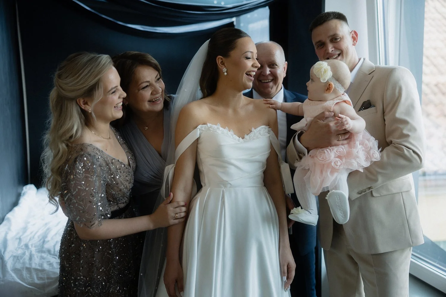 Bride in a white wedding dress smiling with a group of family and friends, including a man holding a baby girl in a pink dress near a window.