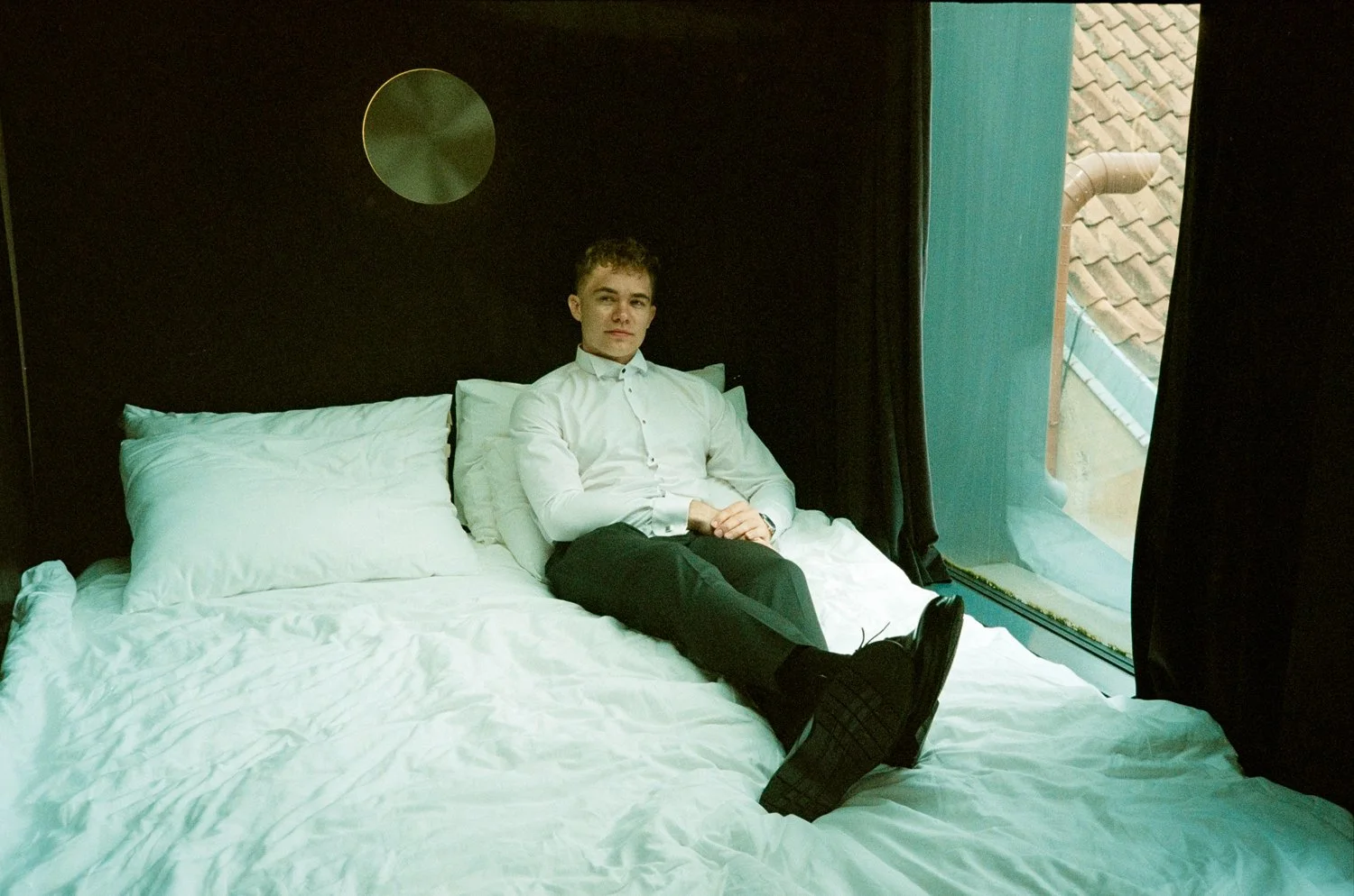 A young man in a white shirt and black pants sitting on a bed in a modern hotel room near a window with a view of rooftops.