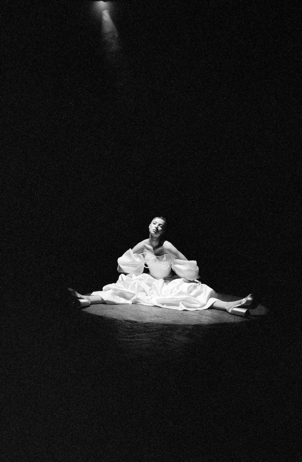 A woman sitting on the floor in a spotlight, wearing a flowing dress and high heels, in a dark room.