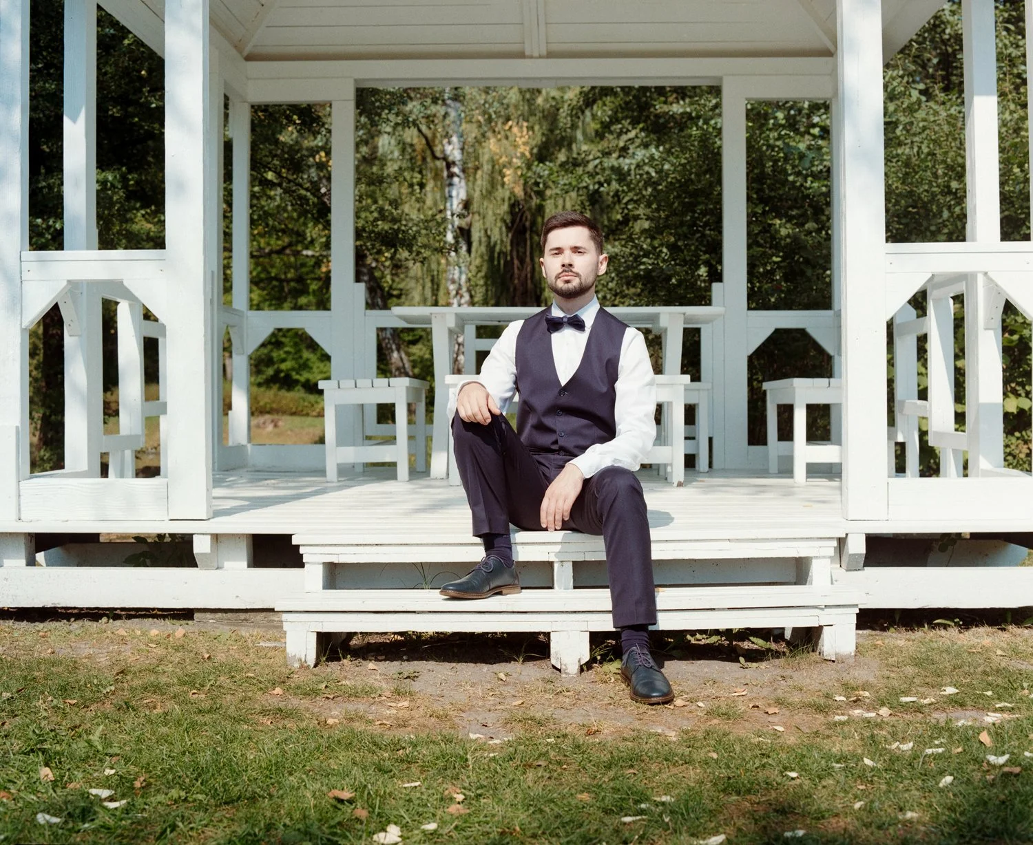 A man in formal attire, wearing a vest and bow tie, sitting on the steps of a white gazebo outdoors, with trees and greenery in the background.