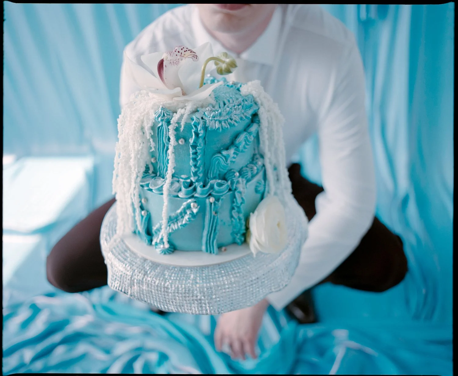 Person in a white shirt holding a tall blue cake with white decorative icing and a white flower on top, standing on a silver cake stand.