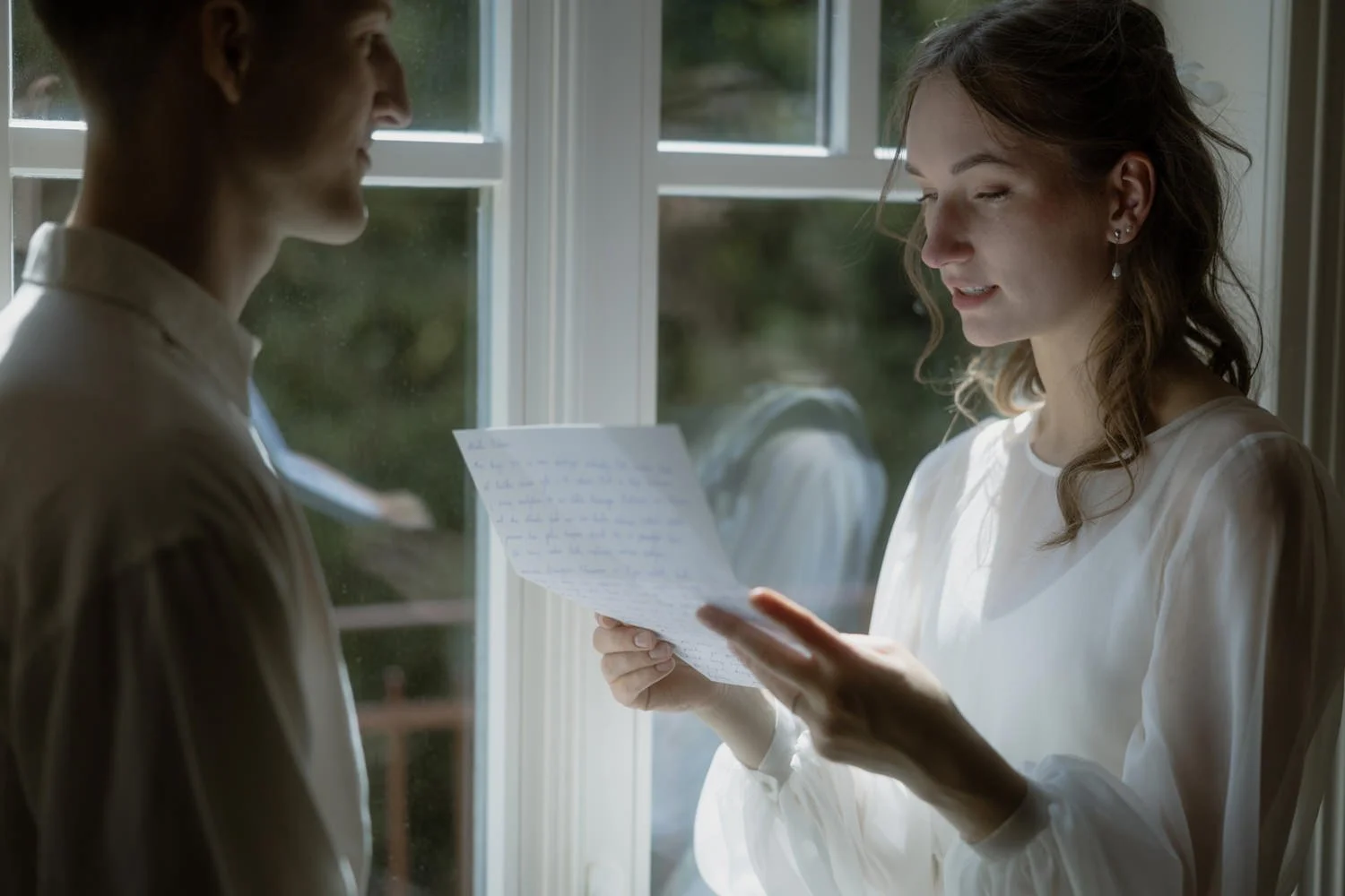 A woman with wavy hair reading a letter in front of a window, with sunlight coming in.