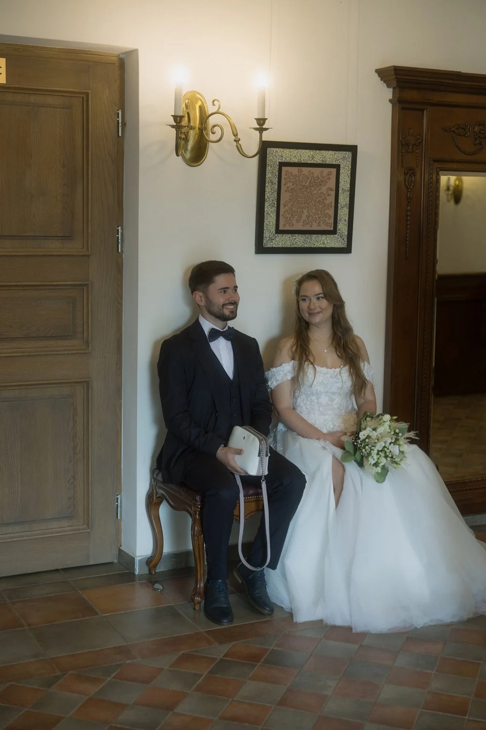 A bride and groom sitting on a wooden bench in a room with wooden paneling and artwork on the wall, dressed in wedding attire, with the bride holding a bouquet of white flowers.