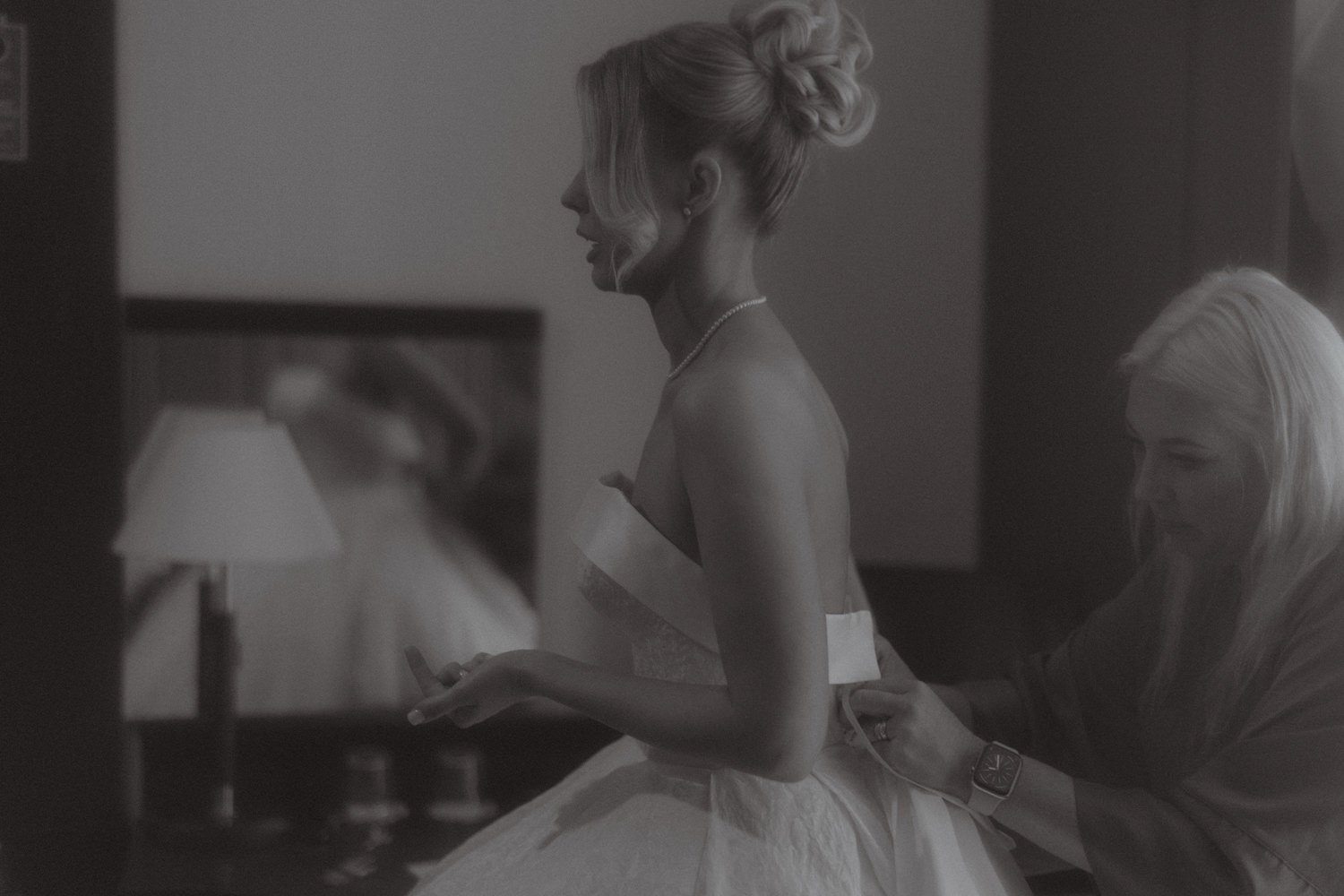 A woman in a wedding dress with her hair styled in an elaborate updo, looking at her phone as another woman assists her, in a room with a small lamp on a nightstand.