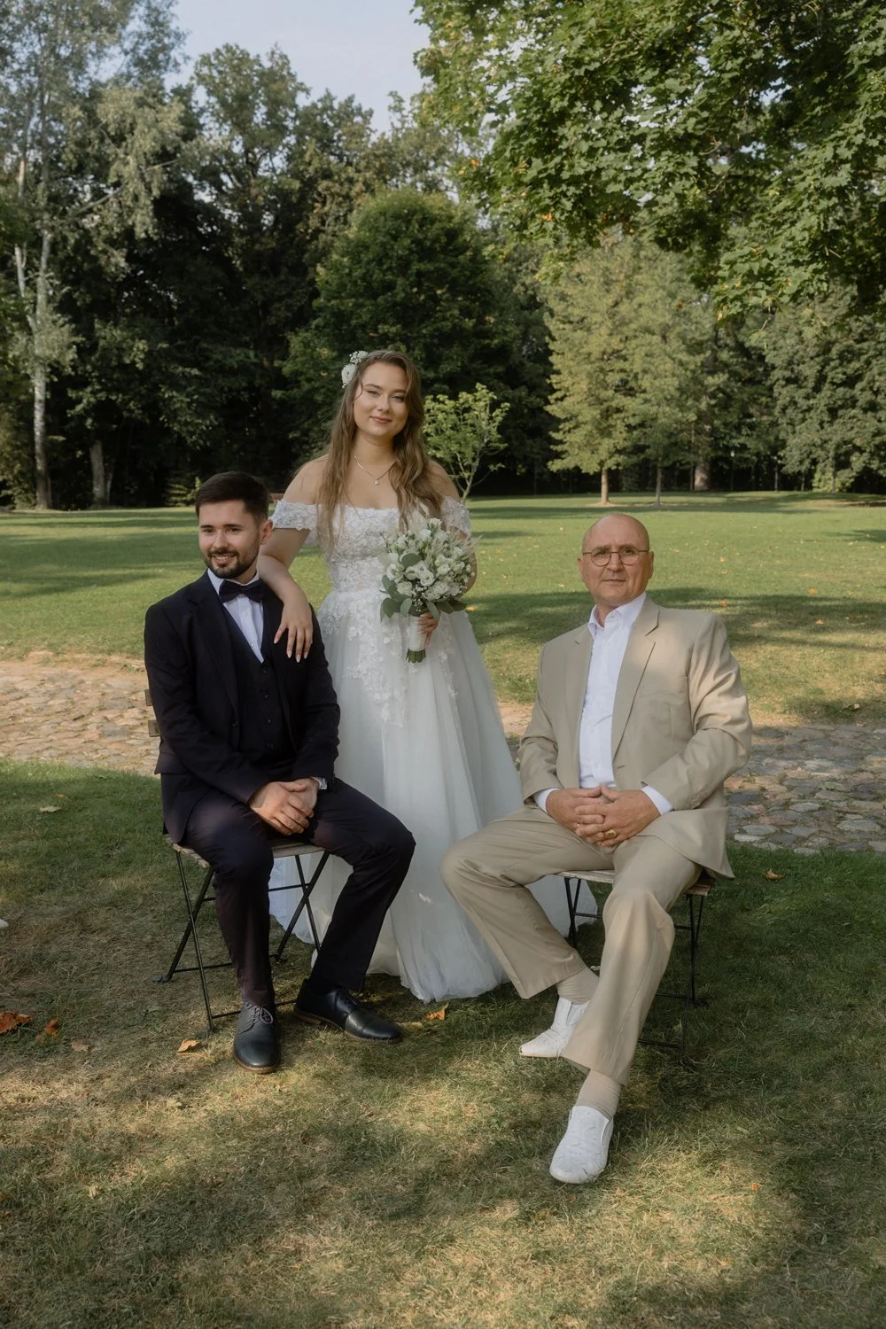 A wedding photo featuring three people outdoors on a grassy area with trees in the background. A woman in a white wedding dress holding a bouquet stands between two men seated on chairs, one in a black tuxedo and the other in a beige suit.
