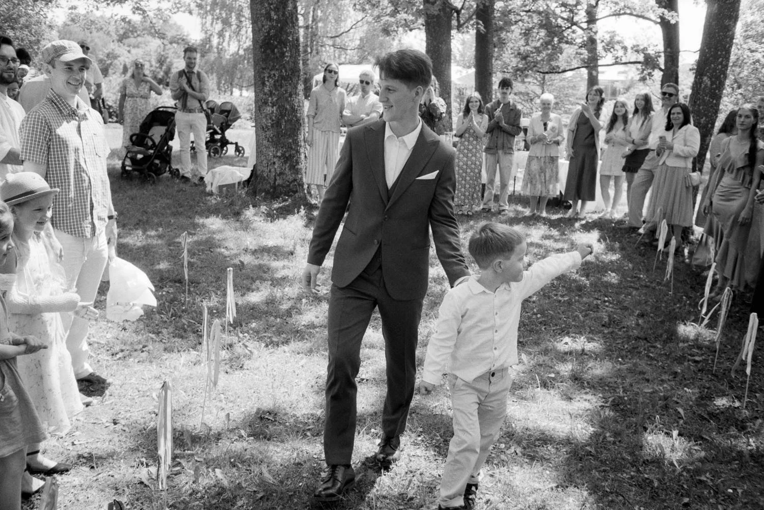 A man in a suit and a young boy are walking through a circle of people outdoors, with children and adults watching and smiling. The scene appears to be a joyful outdoor celebration or event in a park.