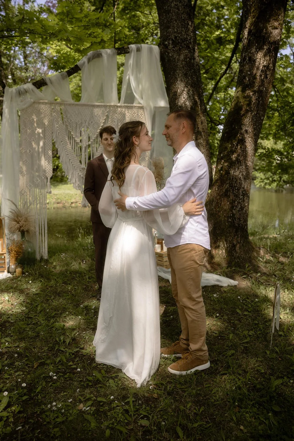 A couple is getting married outdoors by a lake, holding hands, with a officiant in the background. The scene is surrounded by green trees and a decorated canopy.