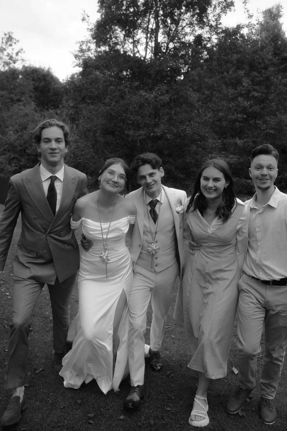 A group of five young adults dressed in formal attire, standing outdoors on grass with trees in the background, smiling at the camera.
