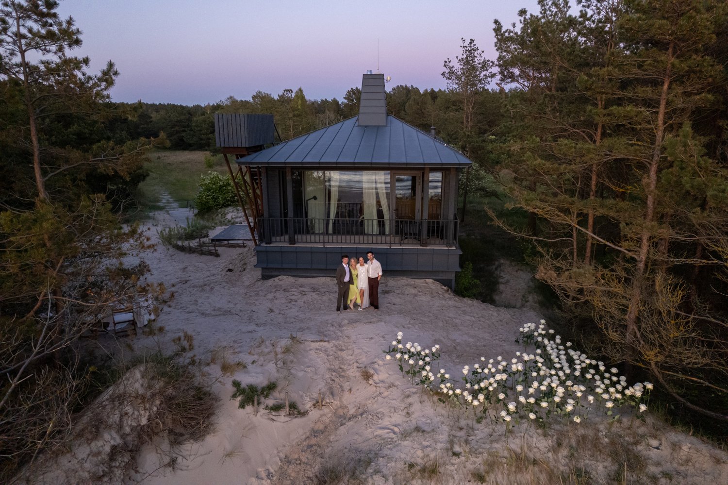 A group of four people standing outside a modern glass house on a sandy area near trees during sunset.
