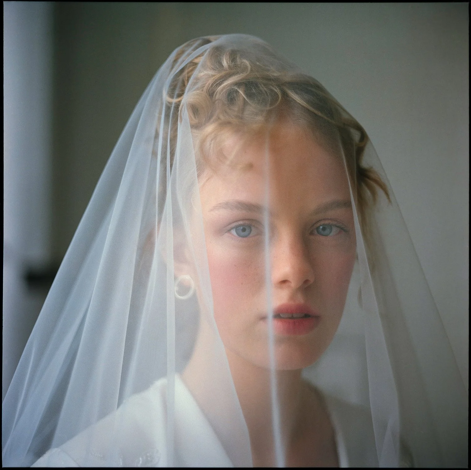 A young bride with short, wavy blonde hair wearing a white veil and a white outfit, looking into the camera.