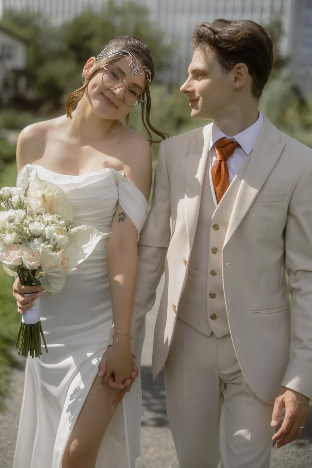 A bride and groom holding hands, smiling, outside on their wedding day, with the bride wearing a white off-shoulder gown, glasses, and a bouquet, and the groom in a cream suit and orange tie.