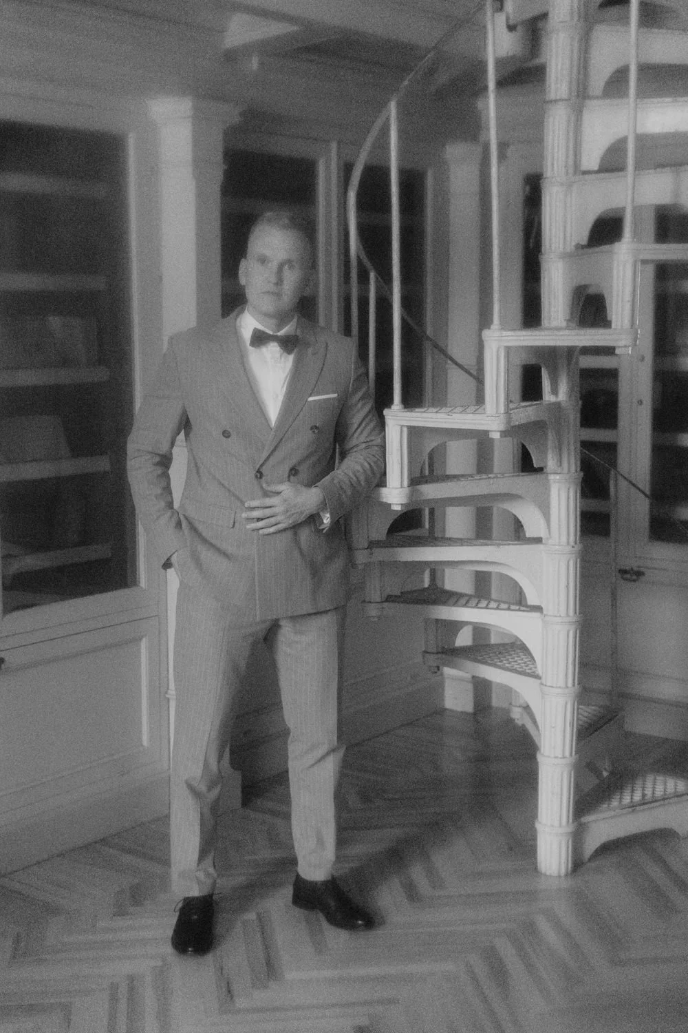 A man in a suit and bow tie standing next to a spiral staircase in a room with wooden flooring and built-in shelves.