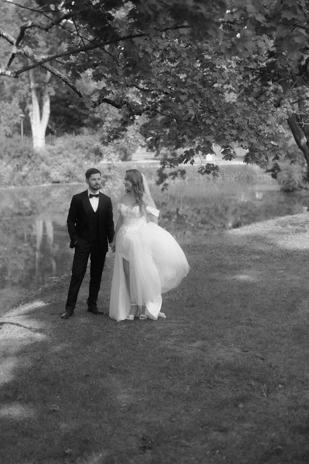 A black and white photograph of a bride and groom standing outdoors near a pond, holding hands and looking at each other. The bride wears a wedding dress with an off-the-shoulder neckline, and the groom is in a suit with a bow tie. They are under a l