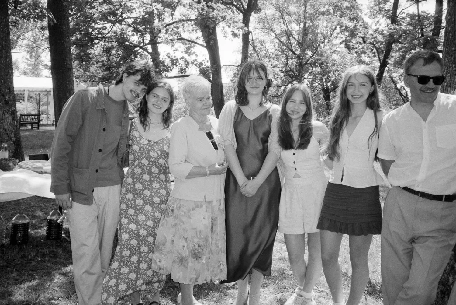 A group of eight people standing outdoors in a park, smiling, with trees and a table with food in the background.