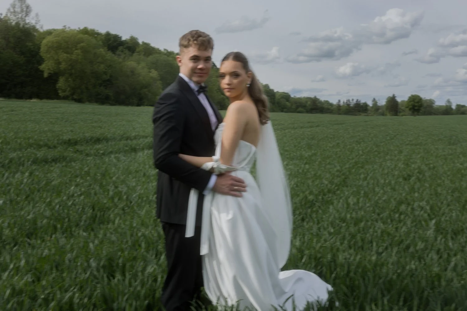 A couple dressed in wedding attire standing in a lush green field with trees and a cloudy sky in the background.