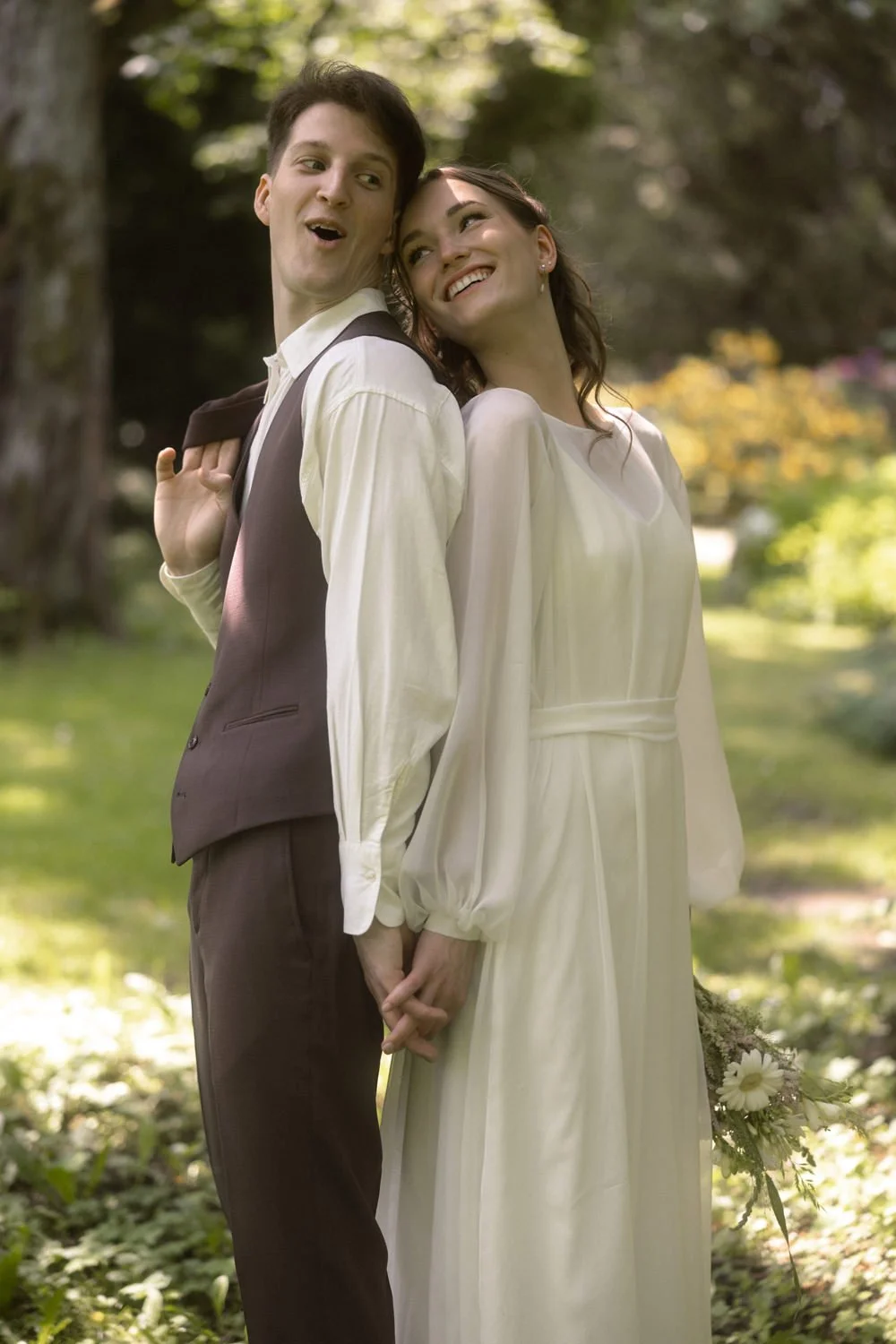 Couple in wedding attire standing outdoors, holding hands back-to-back, smiling, with greenery and trees in the background.