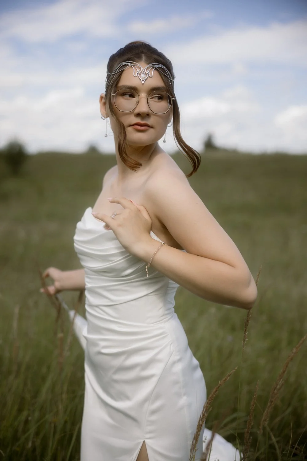 A woman in a white dress standing in a grassy field with hills in the background, looking off to the side with her hand on her shoulder, wearing glasses and jewelry.