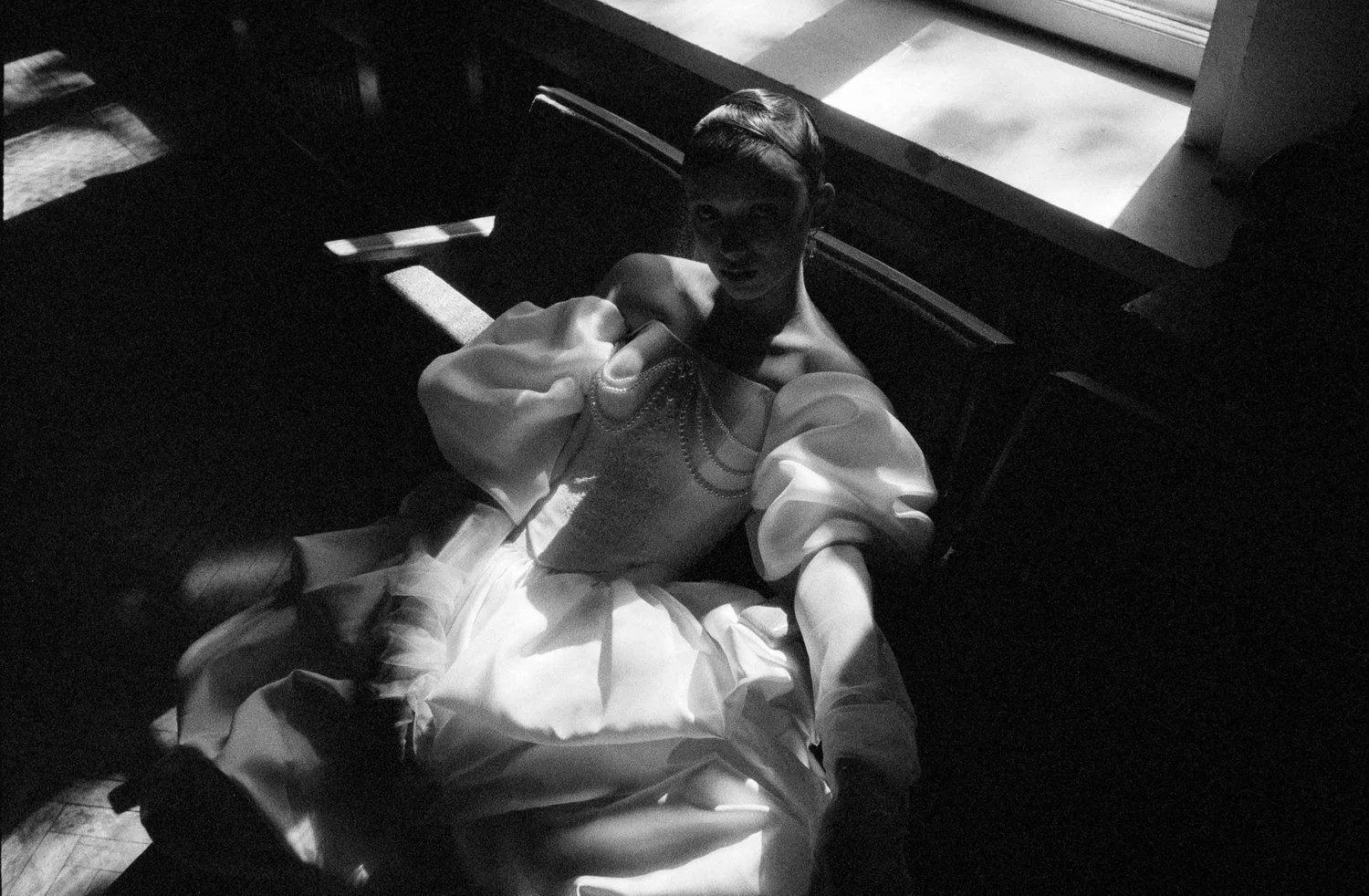 A woman in a vintage dress sitting on a bench near a window with sunlight casting shadows.
