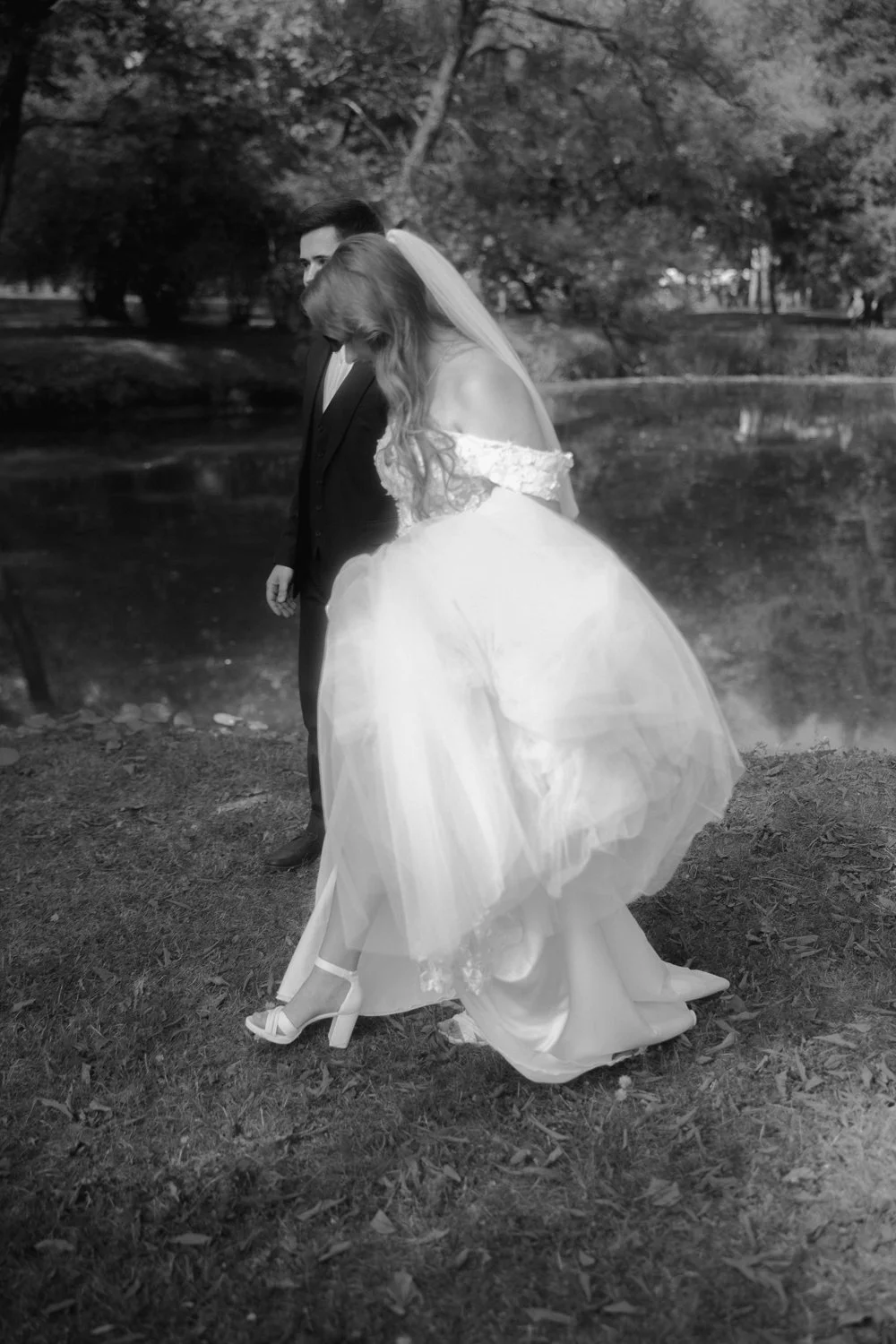 A bride and groom standing by a pond in a park, in a black and white photograph, with the bride wearing a dress with a voluminous skirt and high heels.