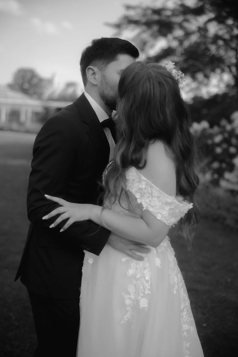 A couple in wedding attire sharing a kiss outdoors, the man in a tuxedo and the woman in a lace dress with floral details, in black and white.
