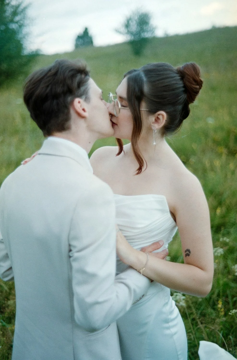 A couple in wedding attire kissing outdoors in a grassy field with trees in the background.