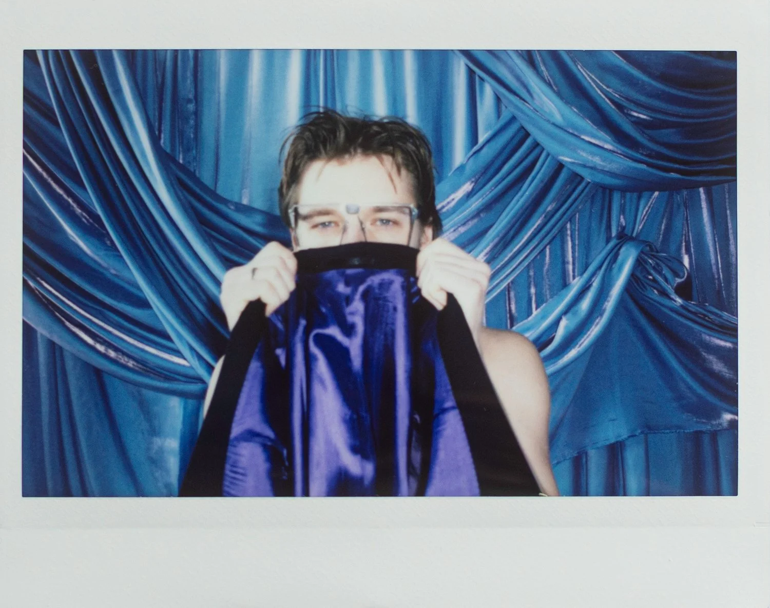Person with short dark hair holding a silky purple and black fabric in front of face, standing in front of blue satin drapes.