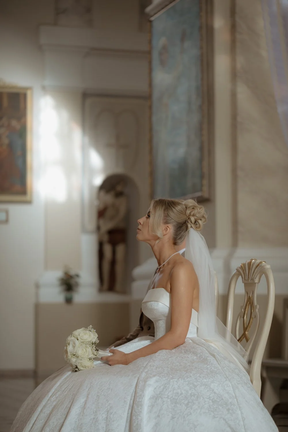 A bride in a white wedding dress sitting in an elegant room holding a bouquet of white roses.