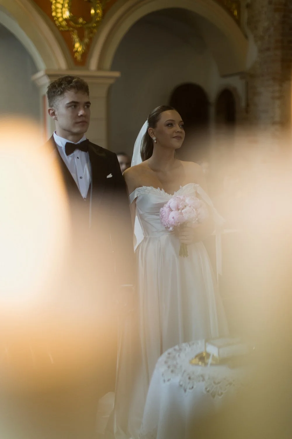 A bride and groom standing during a wedding ceremony inside a church, with the bride holding a bouquet of pink flowers.