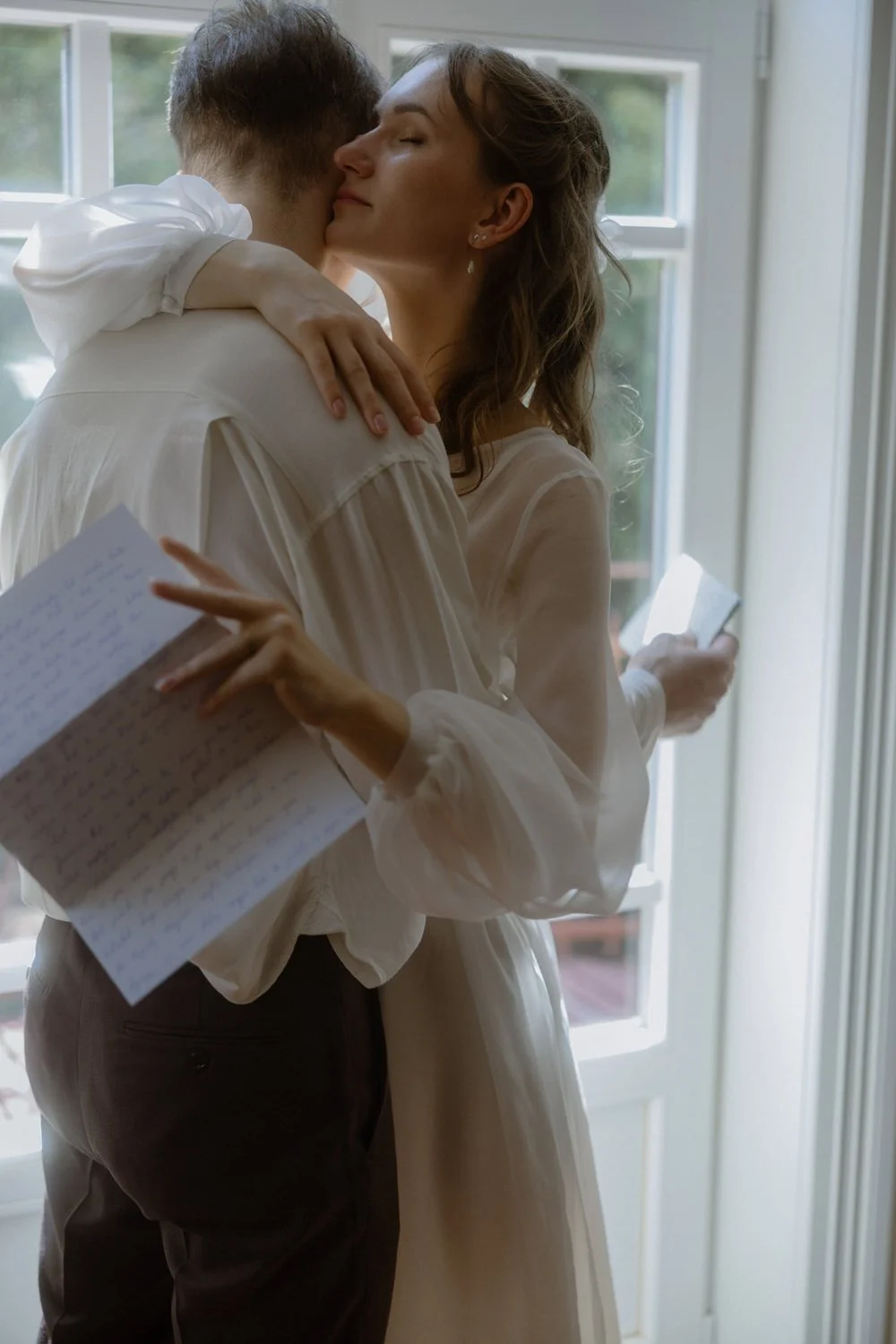 A woman and man hugging inside near a window, with the woman holding a notebook and a phone.