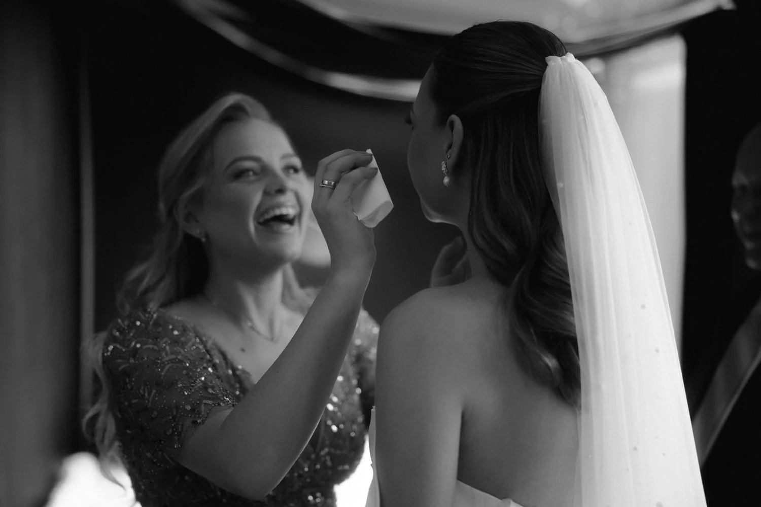 A woman with long hair and pearl earrings wiping the face of a bride with a tissue during her wedding preparation.
