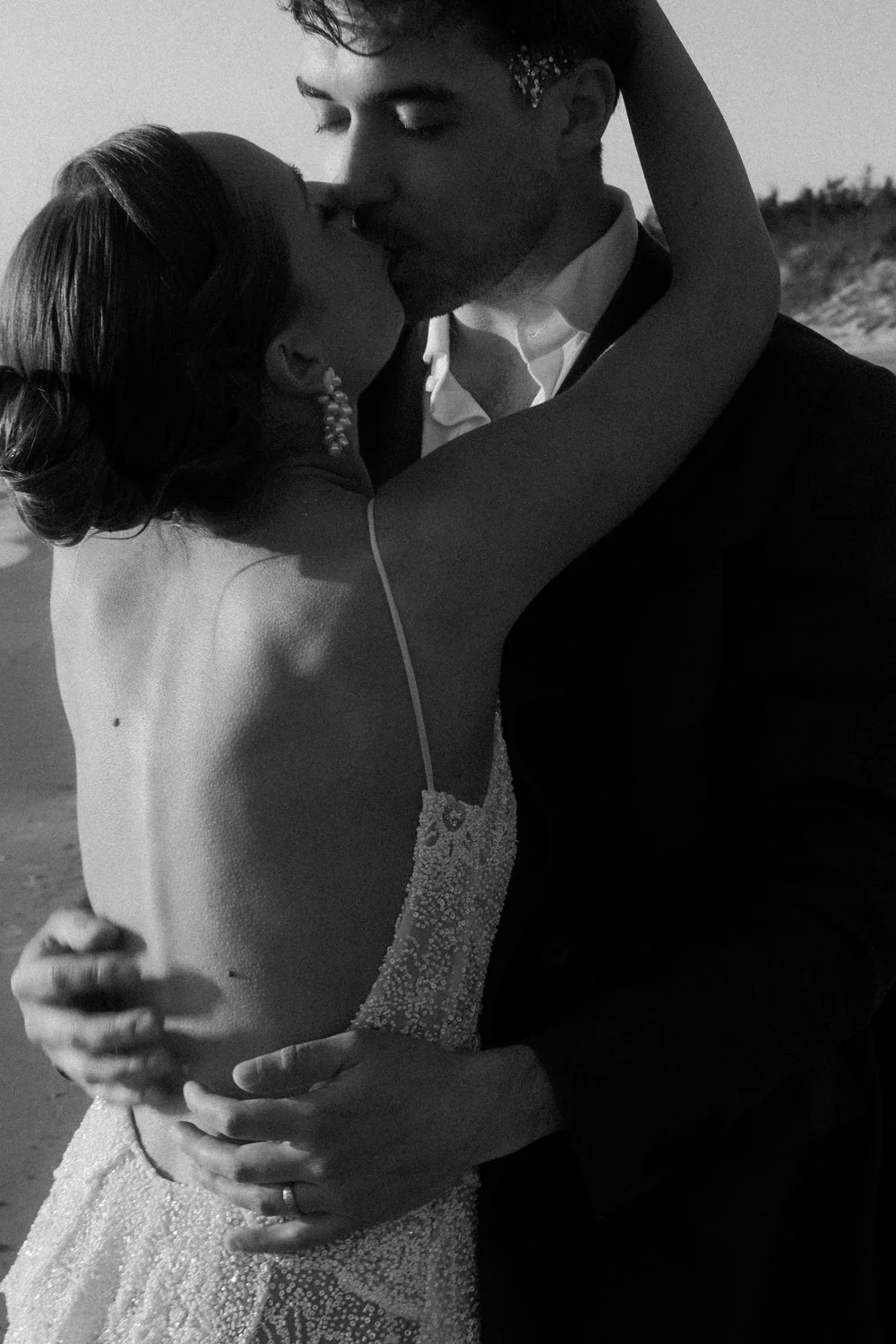 A black and white photo of a couple kissing during their wedding, with the bride's arms around the groom's neck and the groom holding the bride's waist, on a beach or outdoor setting.