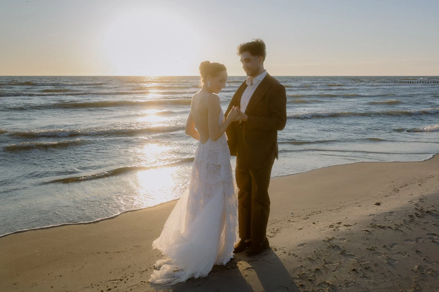 A bride and groom holding hands and smiling on the beach at sunset.