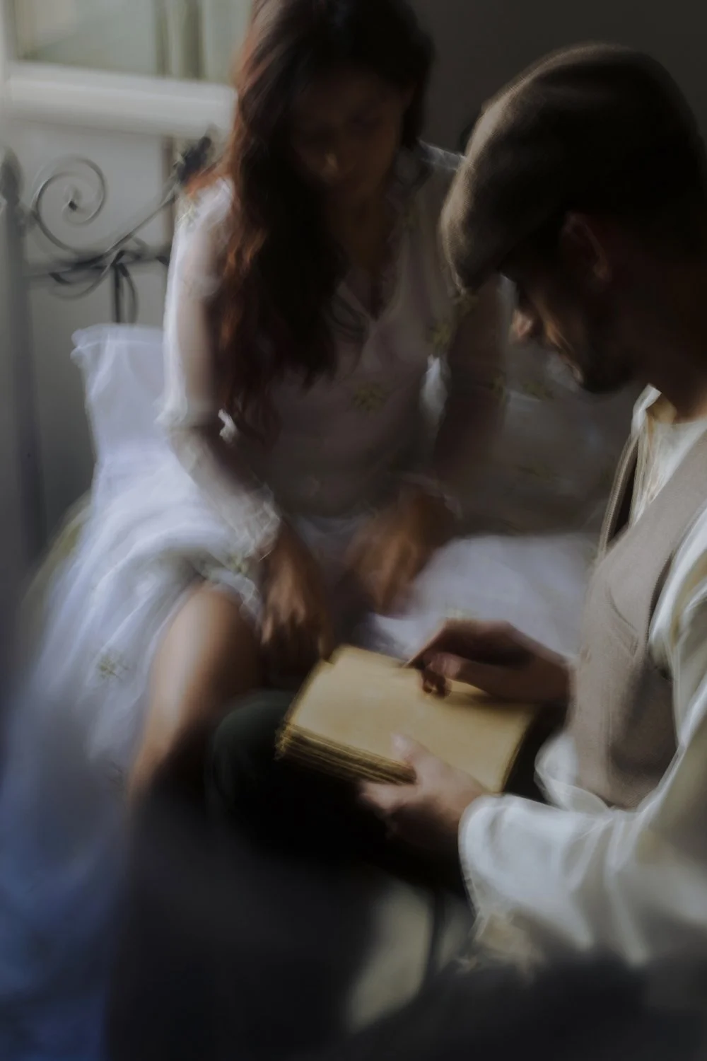 A woman in a white dress - bride , with floral details sitting on a bed, looking at a book held by a man in a vest, with a window in the background.