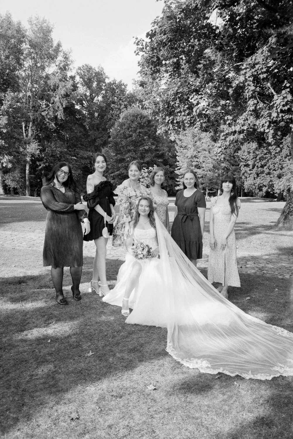 A group of women, including a bride in a wedding dress holding a bouquet, standing outdoors in a park with trees in the background.