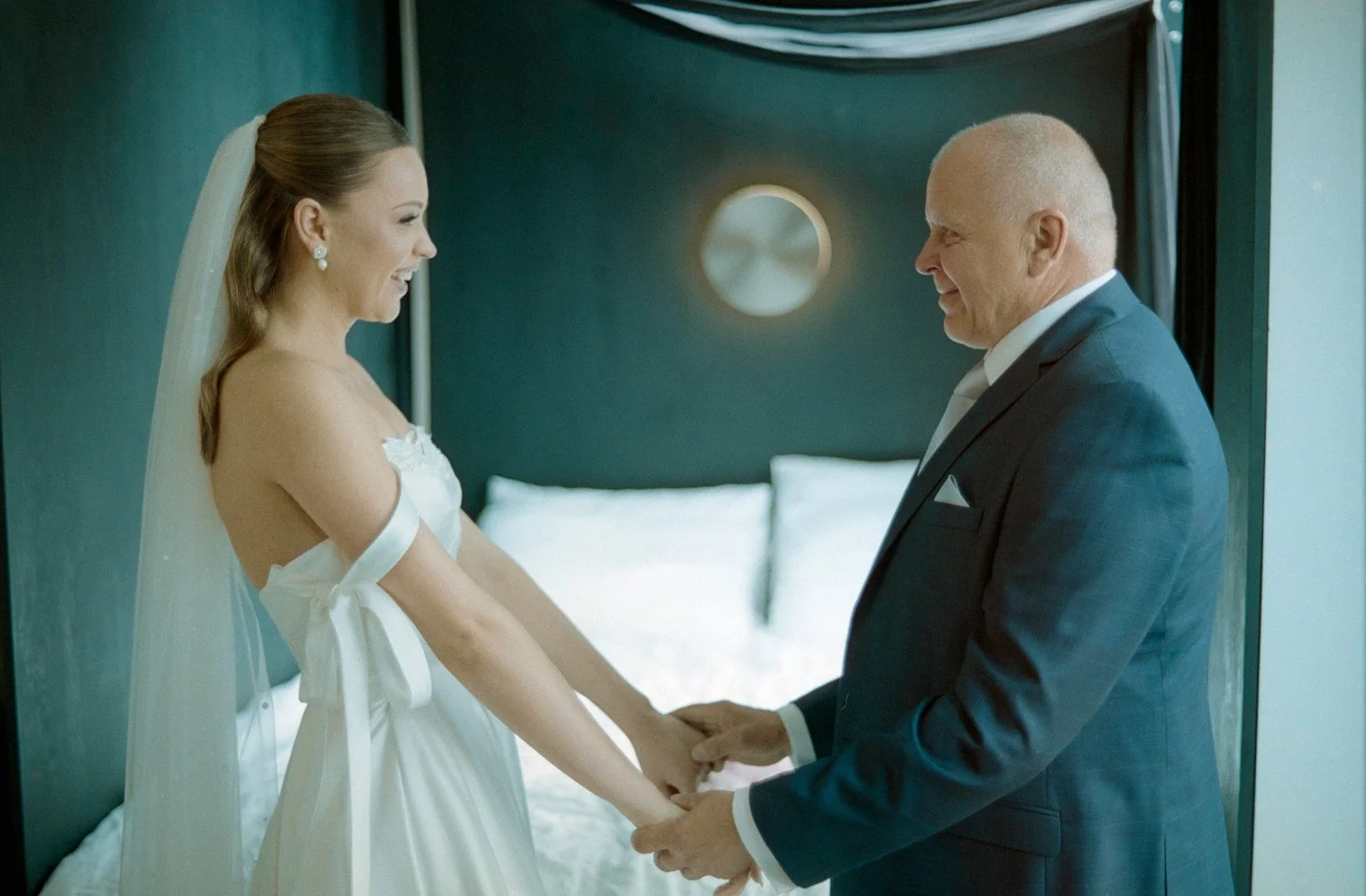 A bride in a white wedding dress and veil holding hands with an older man in a suit, likely her father, during a wedding vows moment in a hotel room.