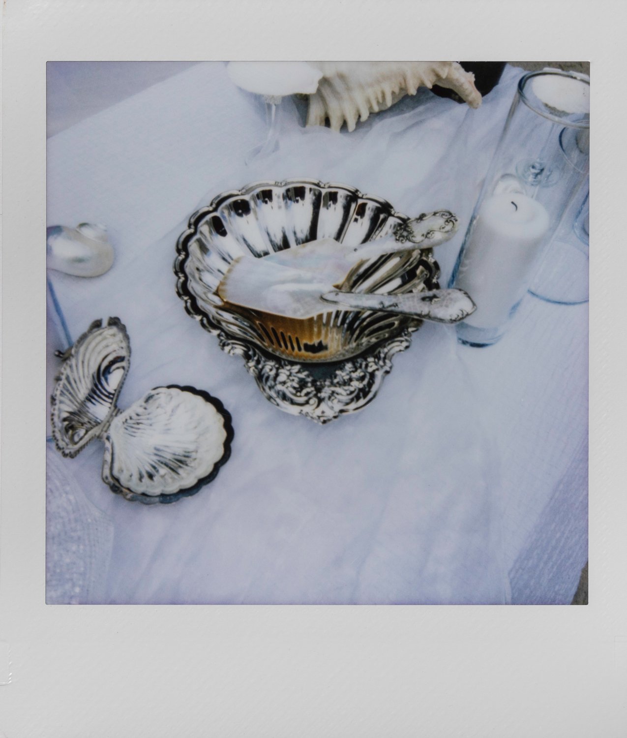 Silver and ceramic dishware, including a bowl, small plates, and utensils, on a white tablecloth with candles and decorative items.