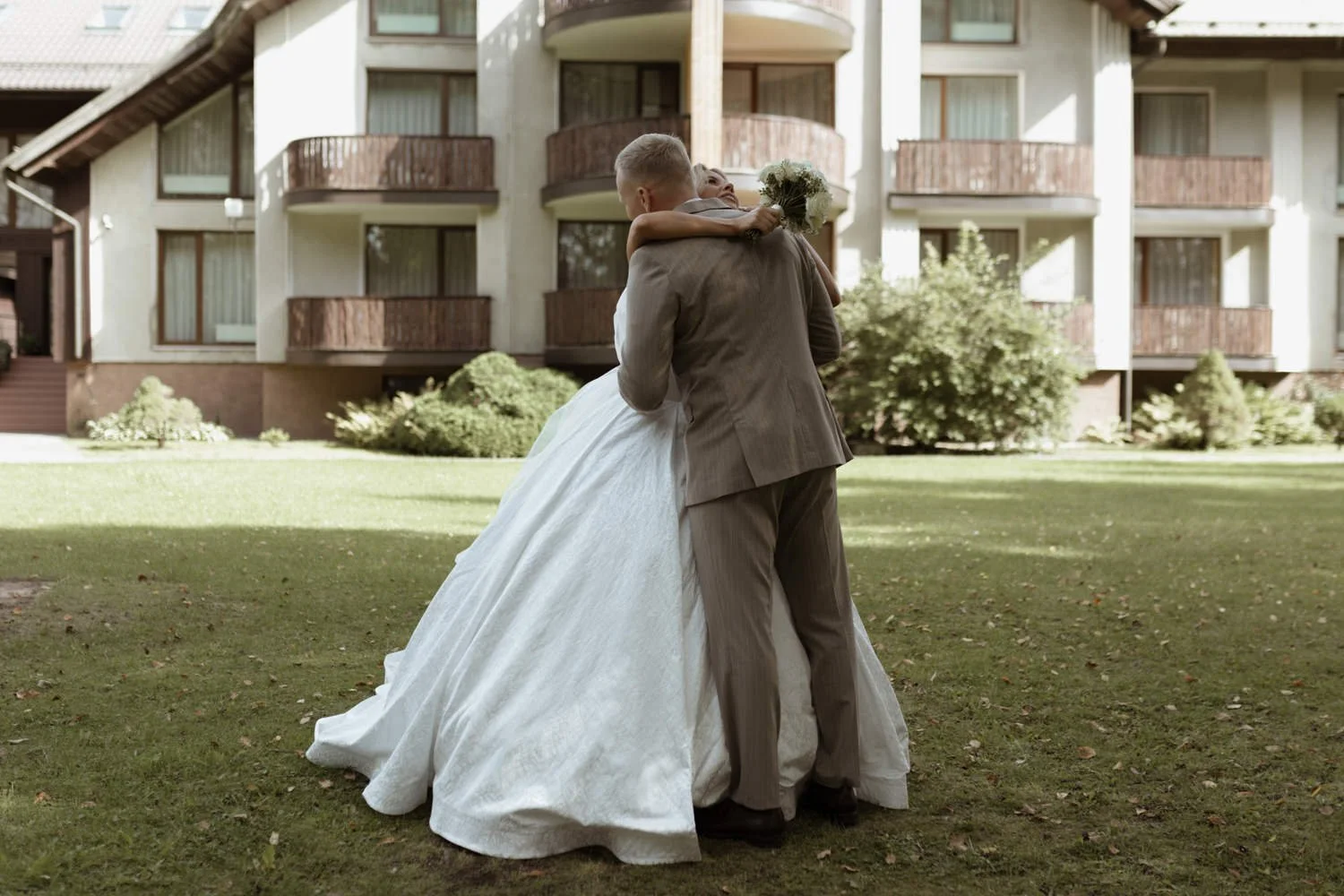 A bride and groom embrace outdoors on a wedding day, with the bride holding a bouquet of flowers, in front of a multi-story residential building.